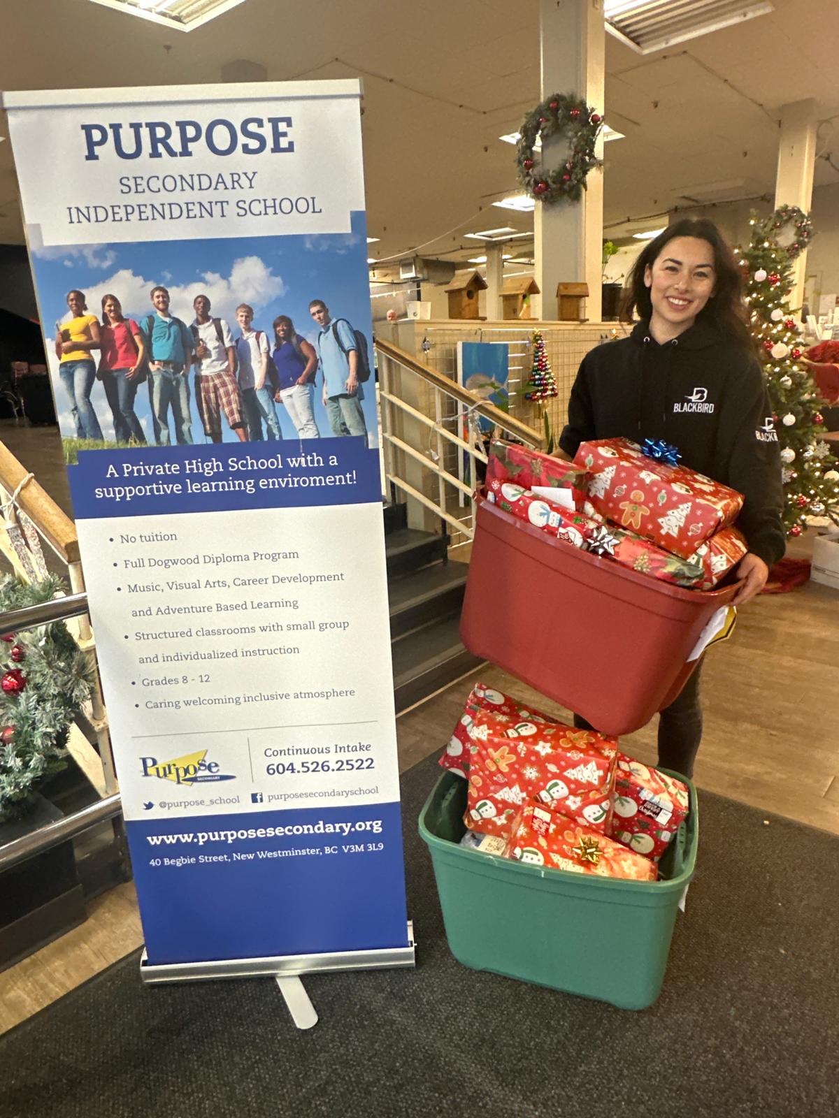 Woman in Blackbird hoodie holding totes of Christmas presents beside a banner of Purpose School