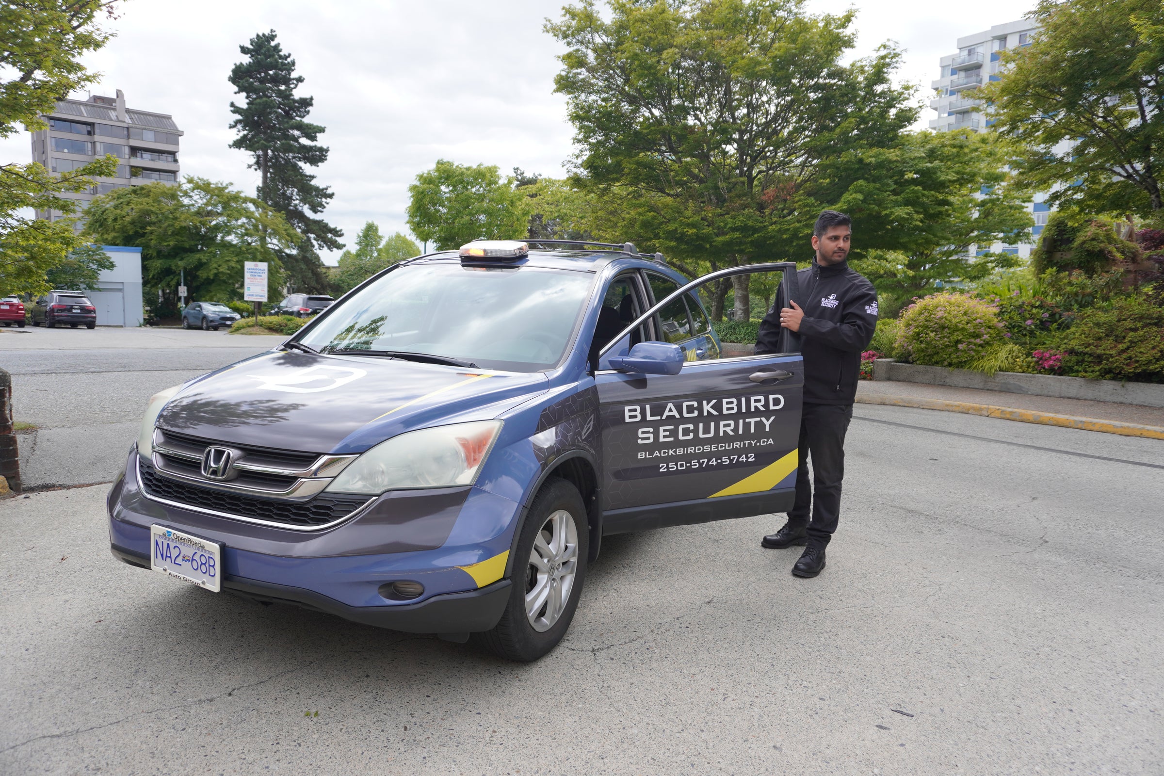 Person standing next to a Blackbird Security vehicle on a street with trees and buildings in the background.