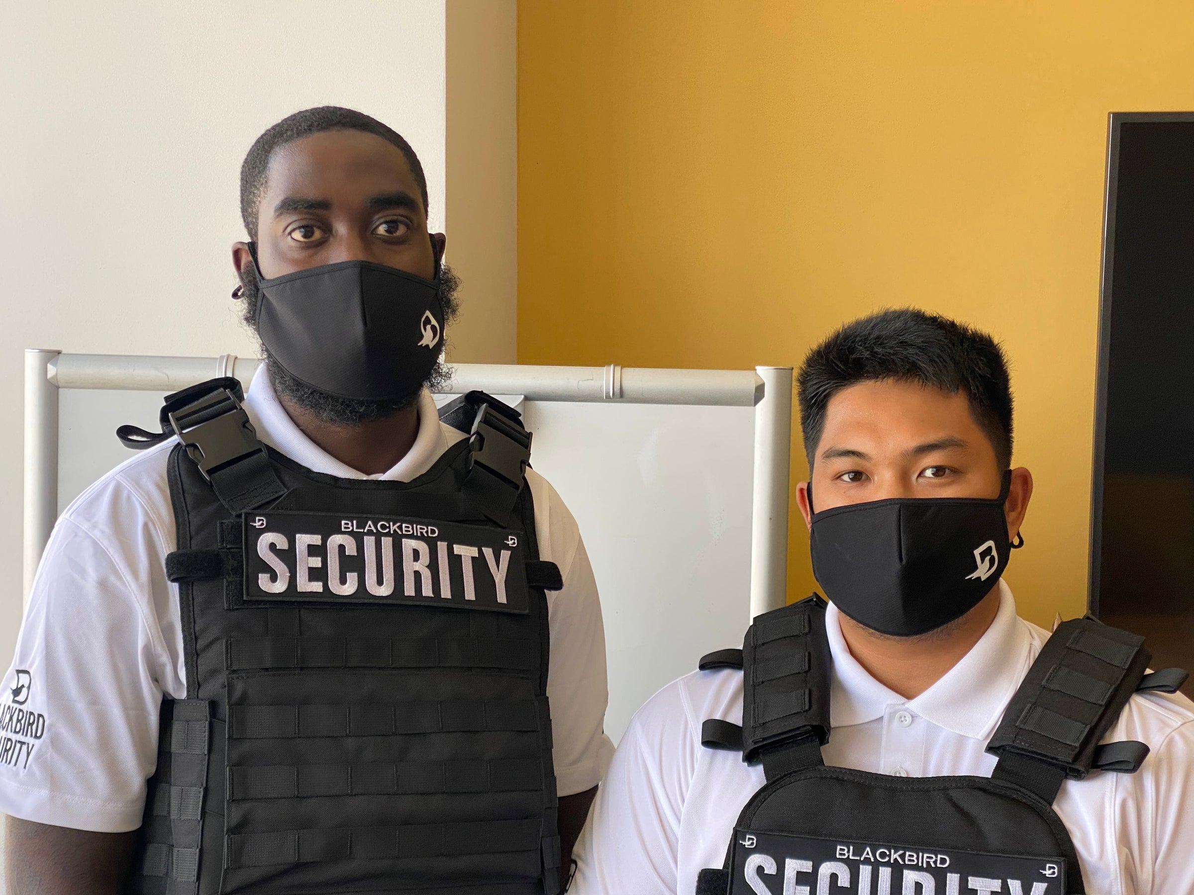 Two security guards wearing Blackbird Security vests and facemasks stand in an office setting.